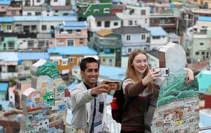 Majid Mushtaq from Pakistan (left) and Elena Kubitzki from Germany have fun taking selfies next to a signature sculpture against the backdrop of colorful houses at the Gamcheon Culture Village in Saha-gu District, Busan, on Oct. 26.